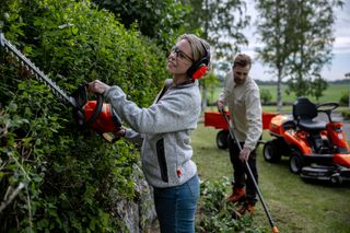 Due persone che tagliano siepi in un giardino, con un trattorino rasaerba sullo sfondo.