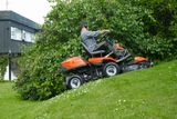A person mowing lawn with a Husqvarna ride-on mower on a grassy slope near a house.