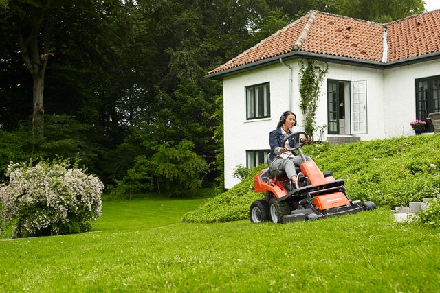 A person mowing lawn with a ride-on mower in a garden with a house in the background.