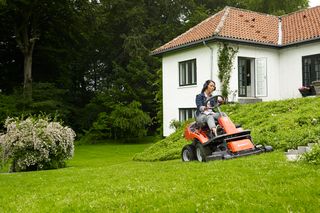 Osoba kosząca trawnik kosiarką samojezdną w ogrodzie z domem w tle.