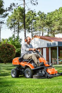 Osoba kosząca trawnik kosiarką samojezdną w ogrodzie z drzewami i kwiatami.