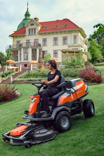 Une personne tond une pelouse avec une tondeuse autoportée devant une grande maison avec jardin.