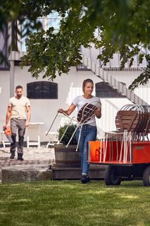 Două persoane care aranjează mobilierul de exterior într-o grădină, cu un cărucior încărcat cu scaune.