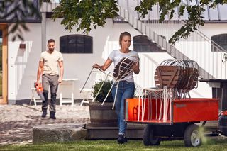 Două persoane care aranjează mobilierul de exterior într-o grădină, cu un cărucior încărcat cu scaune.