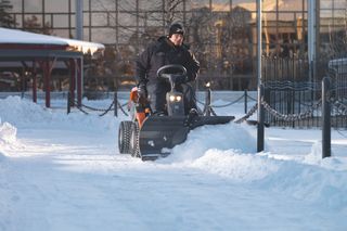 A person operating a snow blower on a snowy path in an urban setting.