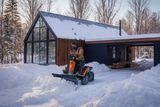 Особа, яка використовує райдер Husqvarna з відвалом для снігу для розчищення засніженої під'їзної дороги біля будинку.