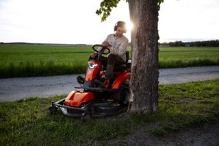 Eine Person, die auf einem Aufsitzmäher sitzt, der unter einem Baum neben einem Feldweg geparkt ist.