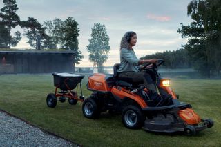 A person operating a ride-on lawn mower with an attached grass spreader in a garden.