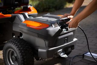 Osoba připojující nabíjecí kabel k prostoru pro akumulátory sekačky Husqvarna se sedící obsluhou.