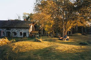 Ein Aufsitzmäher, der auf einer Rasenwiese in der Nähe eines rustikalen Steinhauses geparkt ist, das von Herbstbäumen umgeben ist.
