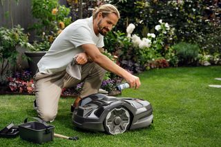 A person kneeling on grass, adjusting settings on a robotic lawn mower in a garden.