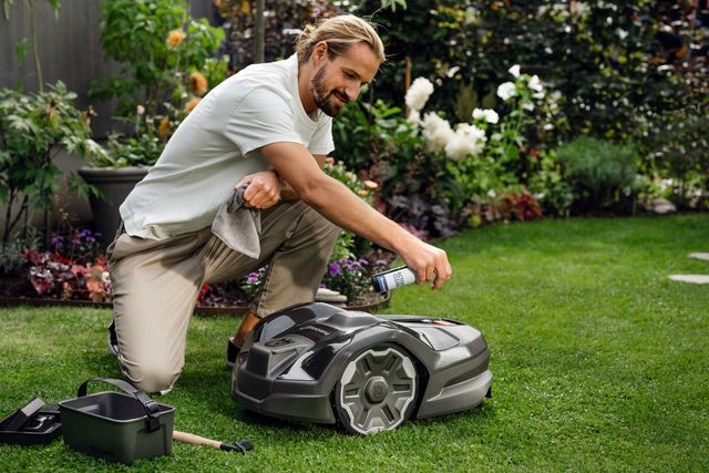 A person kneeling on grass, adjusting settings on a robotic lawn mower in a garden.