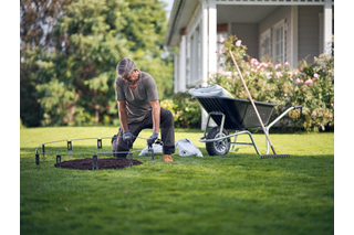 Uma pessoa ajoelhada num relvado, a preparar o solo com ferramentas de jardinagem ao lado de um carrinho de mão.