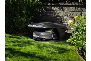 Robotic lawn mower parked in a robotic mower house in a garden near a stone wall.