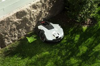 Robotic lawn mower operating on grass near a stone wall and bushes.