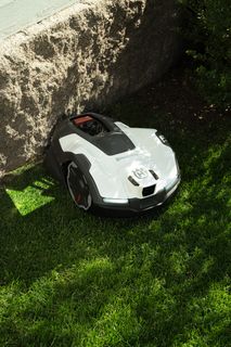 Robotic lawn mower operating on grass near a stone wall and bushes.