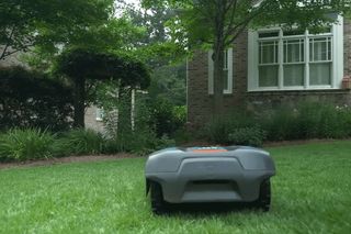 Robotic lawn mower operating in a garden with a brick house in the background.