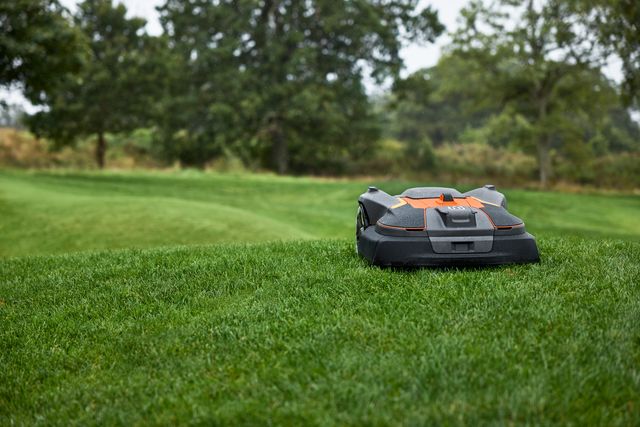 Robotic lawn mower operating on a grassy lawn with trees in the background.