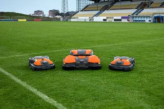 Trois robots tondeuses sur un terrain de football, avec des sièges de stade en arrière-plan.