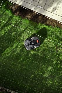 Robotic lawn mower operating on a grass lawn with grid lines overlay, next to a wheelbarrow with soil.