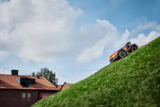 Robotgräsklippare som kör på en brant gräsbevuxen sluttning med hus i bakgrunden.