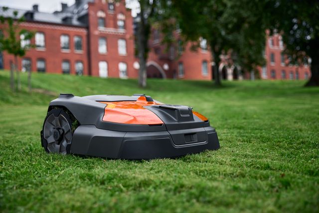 Robotic lawn mower operating on a grassy lawn with red brick buildings in the background.