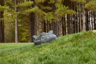 Robô corta-relva a funcionar num declive com relva perto de uma floresta.