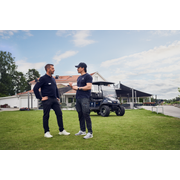 Two people standing on a lawn discussing, with a utility vehicle in the background.