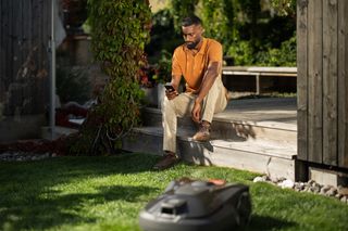 A robotic lawn mower operating on a grassy lawn, with a person sitting in the background.