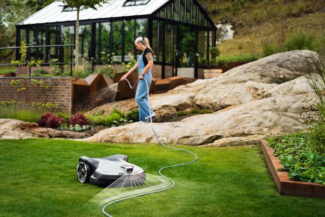 A robotic lawn mower operating in a garden with a person in the background near a greenhouse.