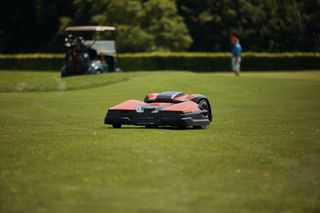 Robot tagliaerba in funzione su un campo da golf con un carrello da golf e un giocatore sullo sfondo.