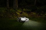 Robotic lawn mower operating in a dimly lit garden with laser guidance lines visible.