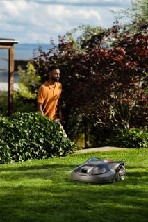 Robotic lawn mower operating on a well-maintained lawn in a garden setting. House and person in the background.