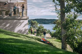 A robotic lawn mower operating on a grassy slope near a lake, with people standing on a balcony and in the garden.