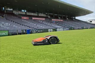 Robotic lawn mower operating on a stadium grass field with empty covered grandstand in background.