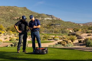 Two people in a desert landscape setting with a robotic lawn mower on the grass.