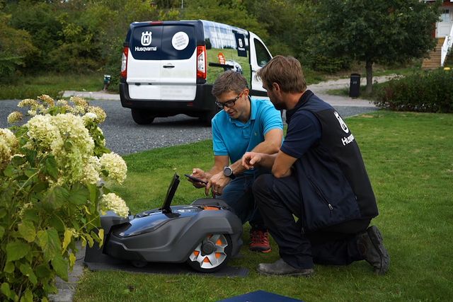 Två personer inspekterar en robotgräsklippare i en trädgård med en Husqvarna-skåpbil i bakgrunden.