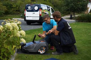 Två personer inspekterar en robotgräsklippare i en trädgård med en Husqvarna-skåpbil i bakgrunden.
