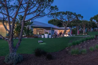 A robotic lawn mower operating on a grassy slope near a modern house at dusk.