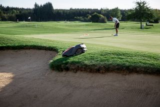 Une tondeuse à gazon robotisée fonctionne près d’un bunker de terrain de golf pendant qu’un golfeur se prépare à rouler.