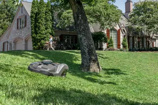 Un robot cortacésped funcionando en una pendiente cubierta de hierba frente a una casa grande, con una persona caminando en el fondo.