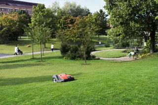Robotska kosilnica deluje v parku, v bližini pa se sprehajajo ljudje.