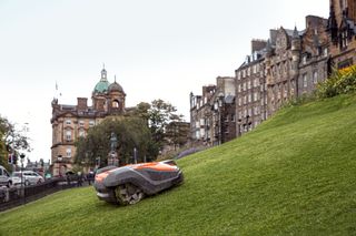 Robotic lawn mower operating on a steep grassy slope in an urban setting.