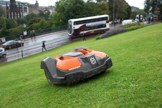 Robotic lawn mower operating on a grassy slope near a busy street with a bus passing by.