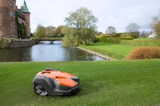 Robô corta-relva que opera num relvado cuidado perto de um castelo e de um lago.