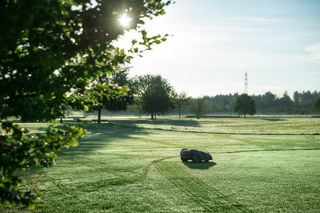 Robotic lawn mower operating on a grassy field with trees in the background.