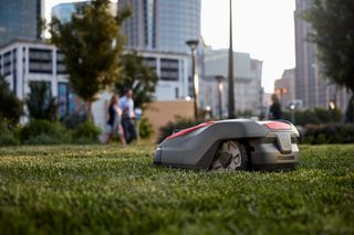 Robotic lawn mower operating in an urban park setting with people walking in the background.