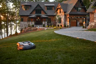 Robotic lawn mower operating on a well-maintained lawn in front of a large house by a lake.
