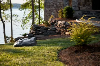 Robotic lawn mower operating on a grassy lawn near a stone building by a lake.