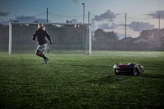 A person playing soccer in the rain while a robotic lawn mower operates on the field.